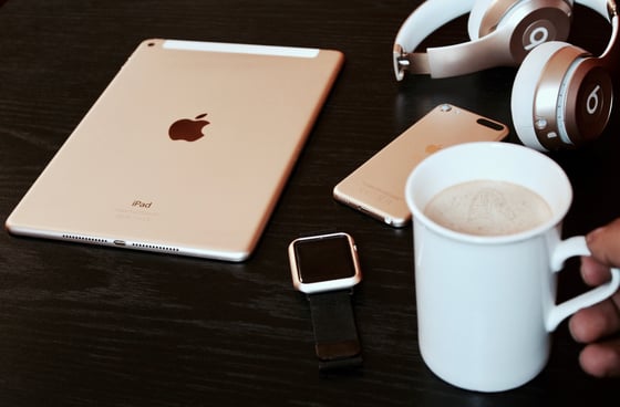 An Apple iPad, watch, and iPhone sit on a table beside Beats headphones and a cup of coffee